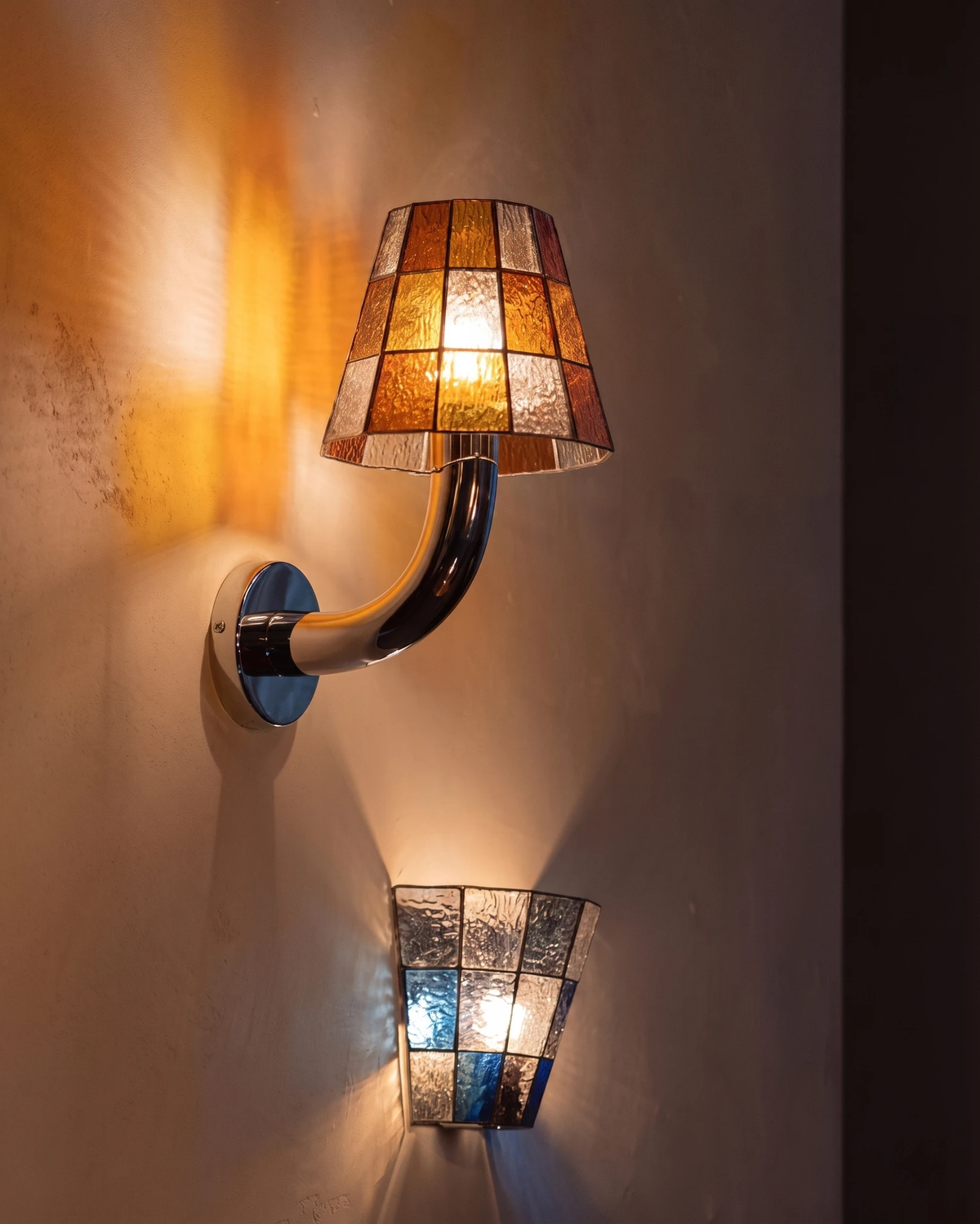 Stained glass wall sconce TESSERA No.01 in amber glowing on a warm plaster wall above a smaller blue mosaic wall light