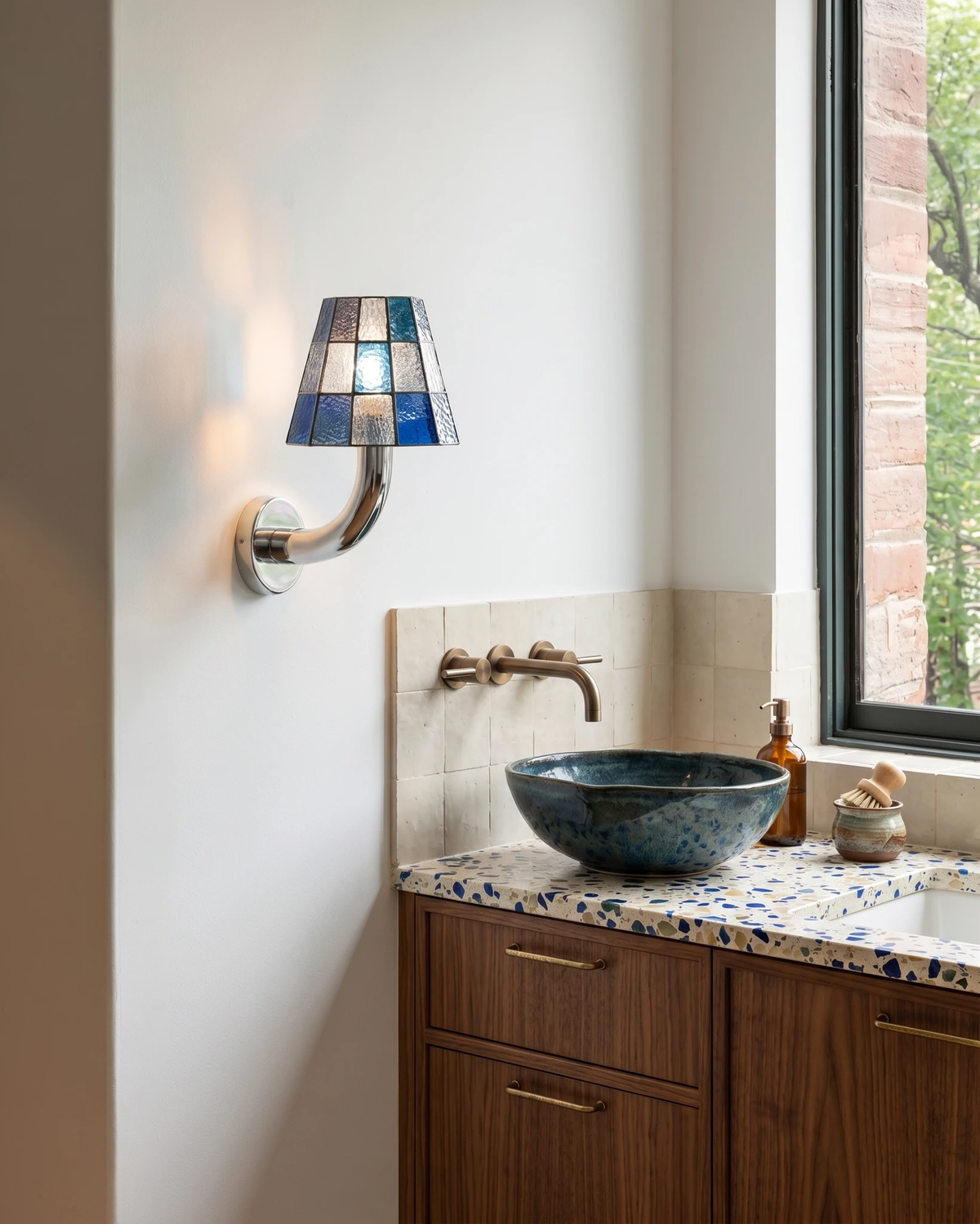 Stained glass wall sconce TESSERA No.01 in blue lit beside a walnut vanity with a blue vessel sink and terrazzo top