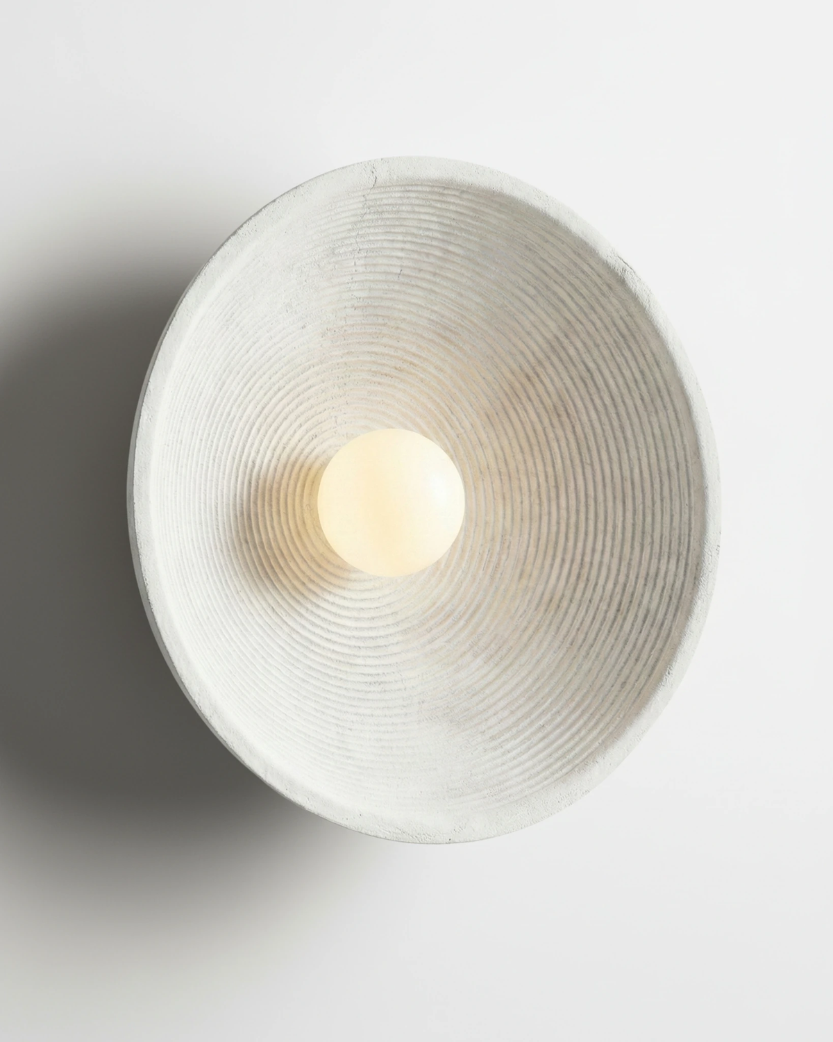 Stone-look wall sconce ECCO No.01 in warm cream, concentric ridged bowl with a glowing pearl bulb, switched on