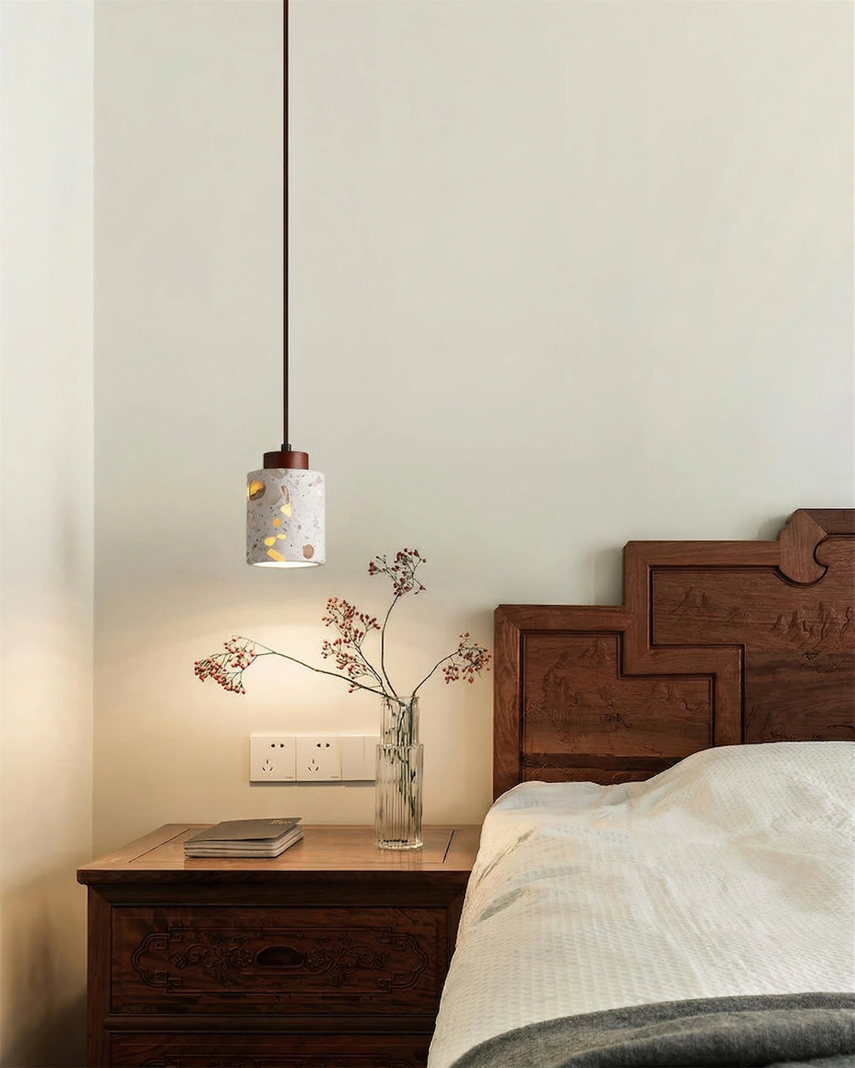 Terrazzo pendant light GROTTA No.01 cylinder lit beside a carved wooden headboard and a nightstand with flowers