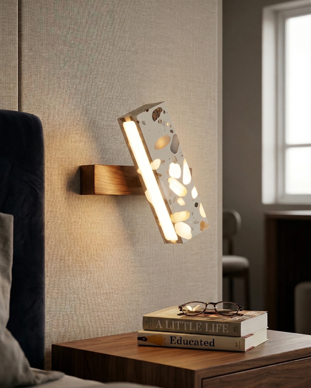 Terrazzo wall sconce PIETRA No.07 switched on beside a blue headboard above a nightstand with books