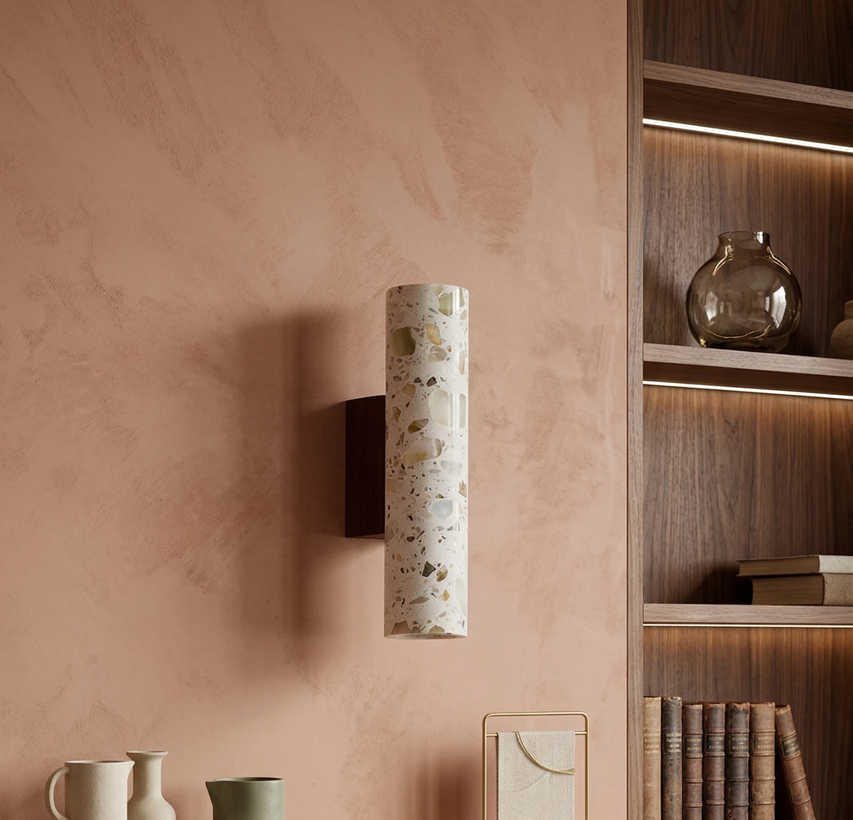 Terrazzo wall sconce INCA No.02 lit on a pink plaster wall beside a wood shelving unit with vases