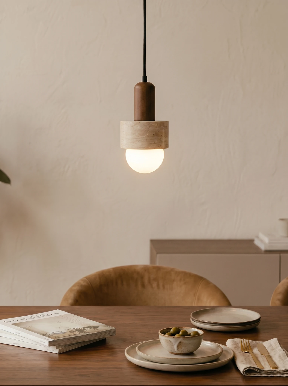 Travertine pendant light TONDO No.05 lit low over a small wood table set with a plate and bowl beside a velvet chair