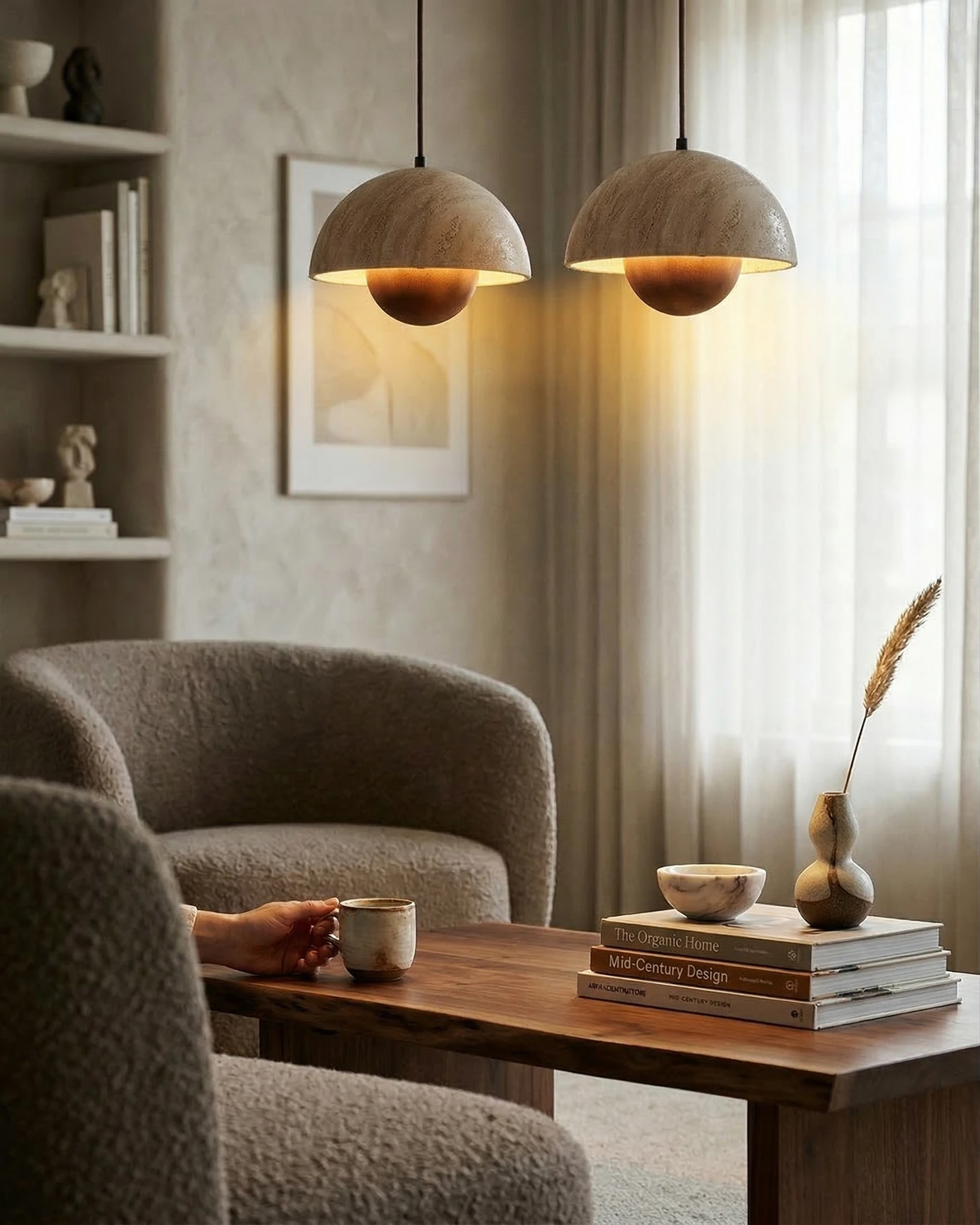 Pair of travertine pendant lights SOLE No.01 lit over a wooden table between boucle armchairs, a hand holding a cup