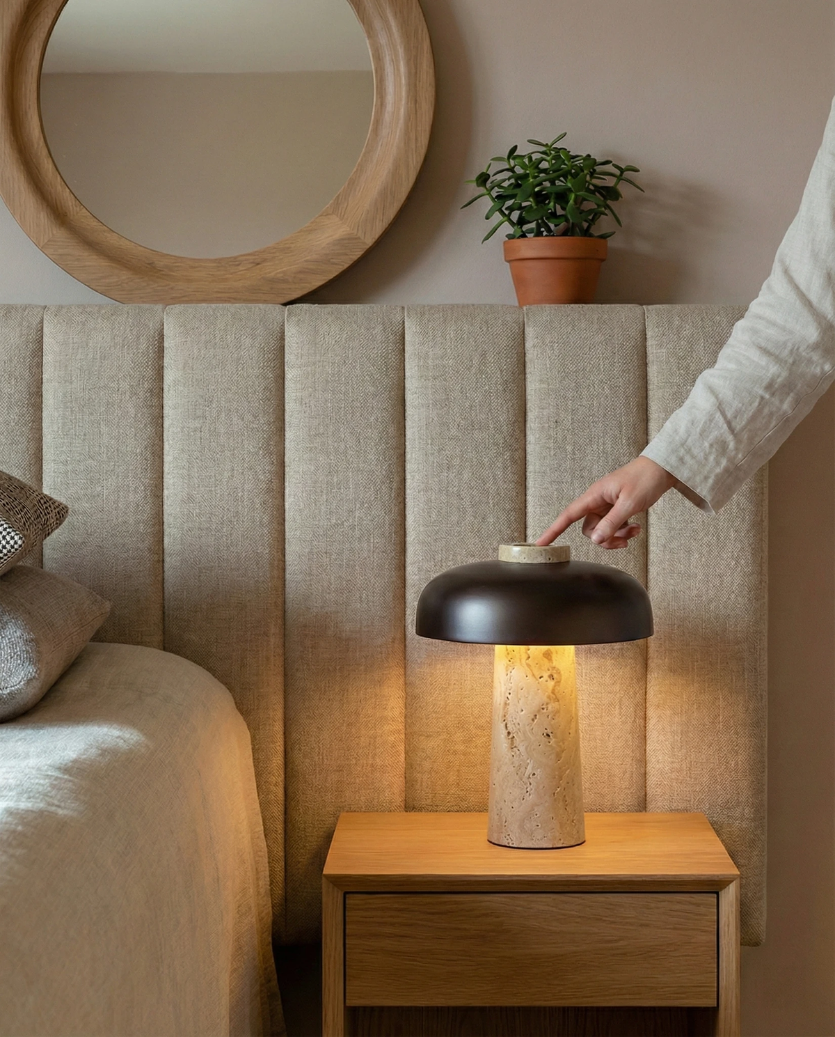 Travertine table lamp ROCCA No.03 lit on an oak nightstand as a hand reaches for it beside a tufted headboard