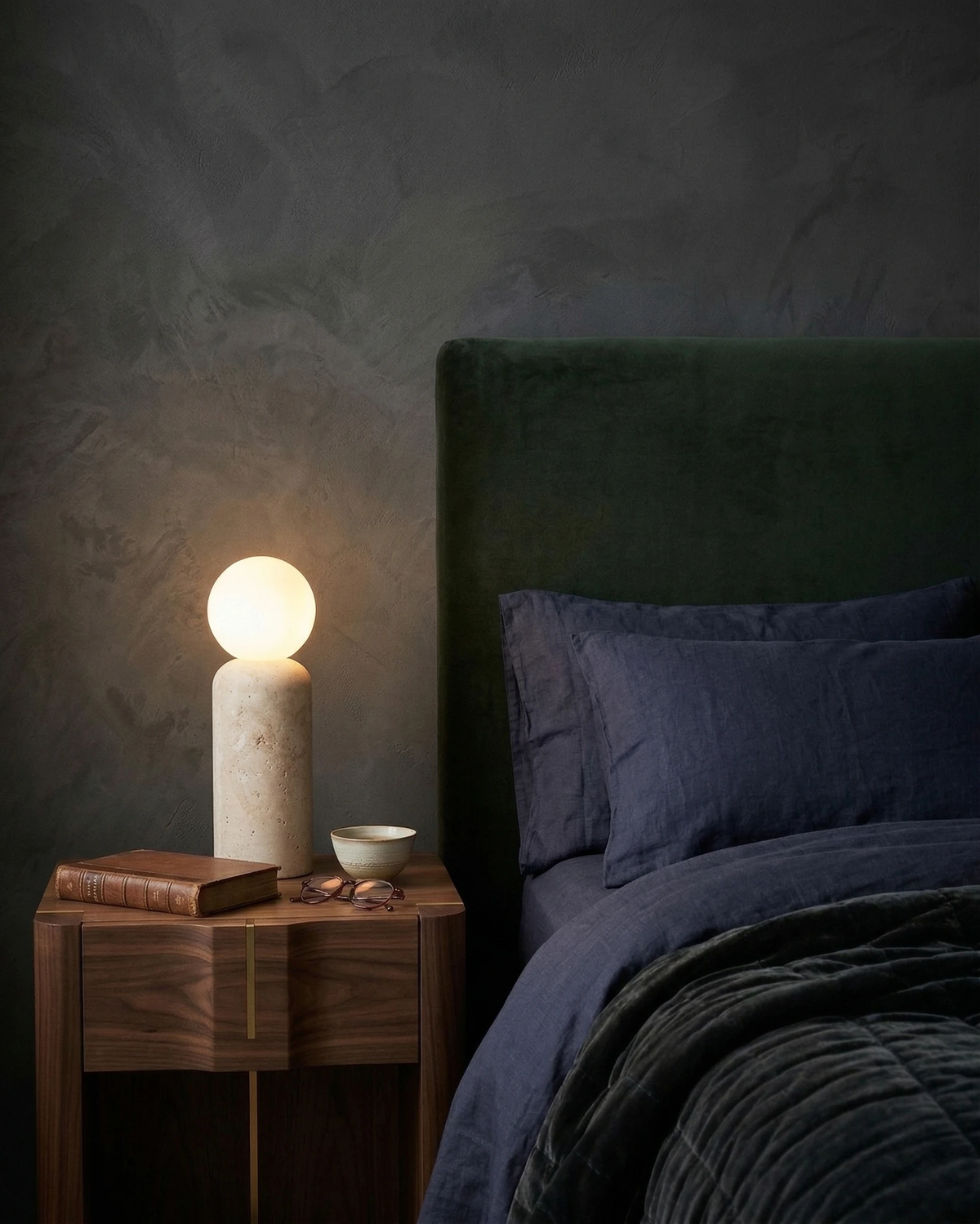 Travertine table lamp MORA No.04 cylindrical plinth lit on a walnut nightstand beside a dark green headboard