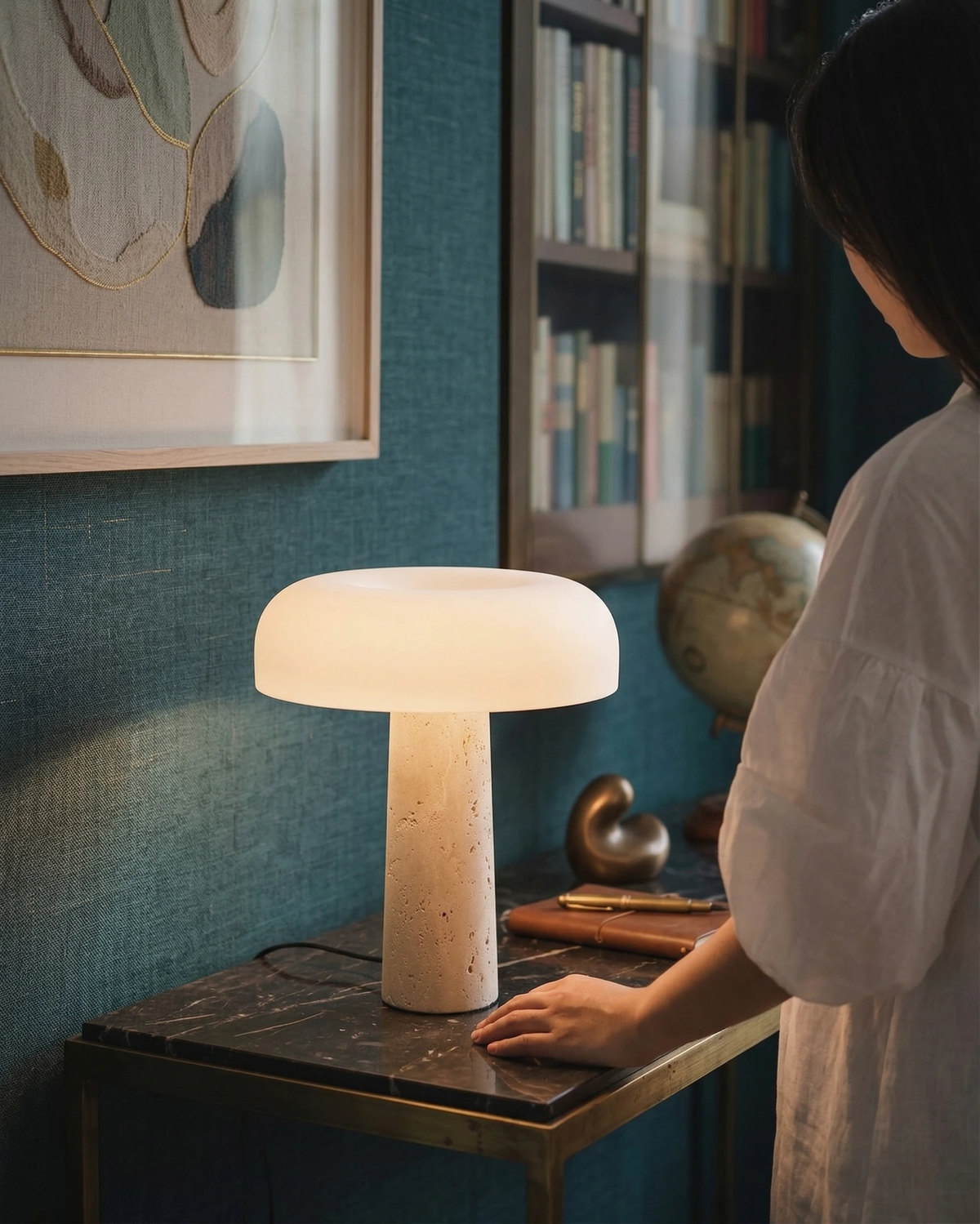 Travertine table lamp MORA No.03 lit on a black marble side table as a woman stands beside it by a bookshelf