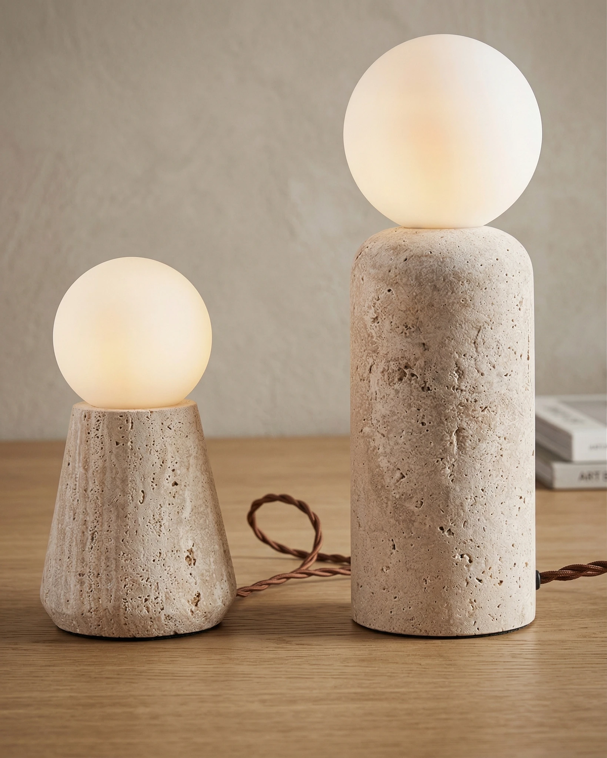 Two travertine table lamps MORA No.04, conical and cylindrical plinths with lit opal globes side by side on a wood table
