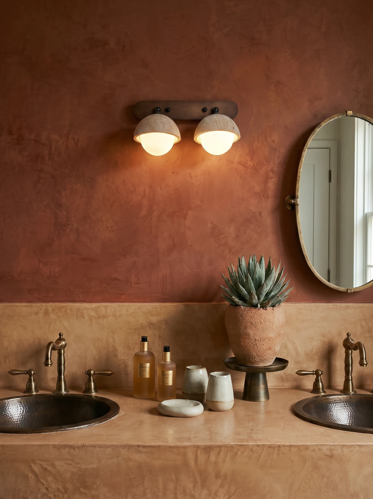 Travertine wall sconce AURA No.02 with two lit globes on a terracotta plaster wall above a double sink vanity