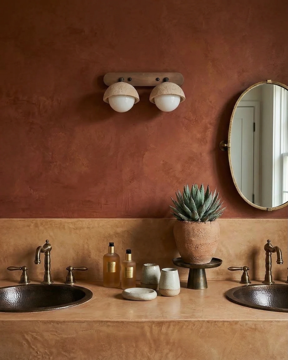 Travertine wall sconce AURA No.02 double globe glowing on a rust plaster wall between an oval mirror and copper sinks