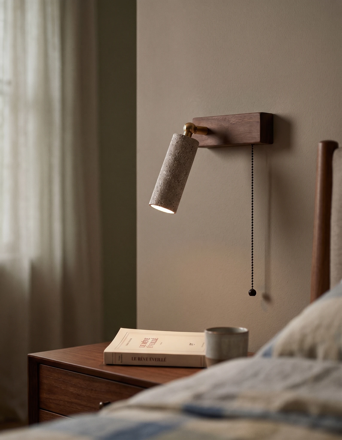 Travertine wall sconce VITRU No.01 with walnut block and pull chain, lit beside a bed over a nightstand with a book