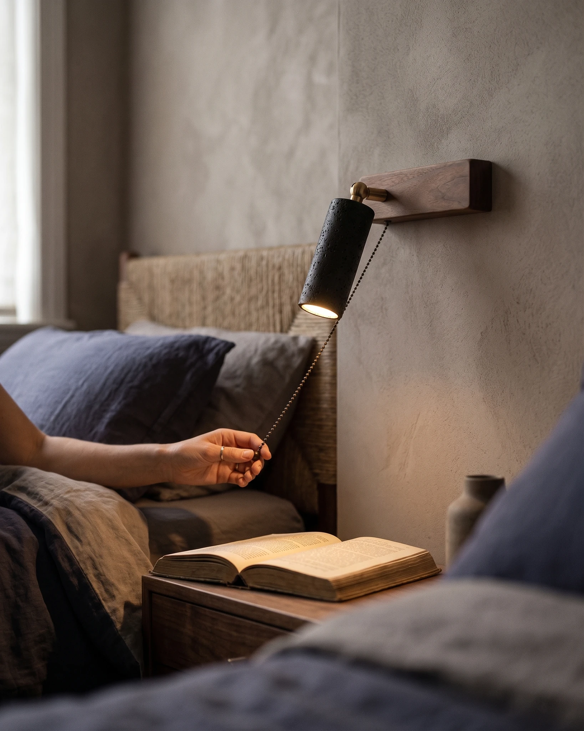 Travertine wall sconce VITRU No.01 in volcano black, lit above a bed where a person lies reading a book