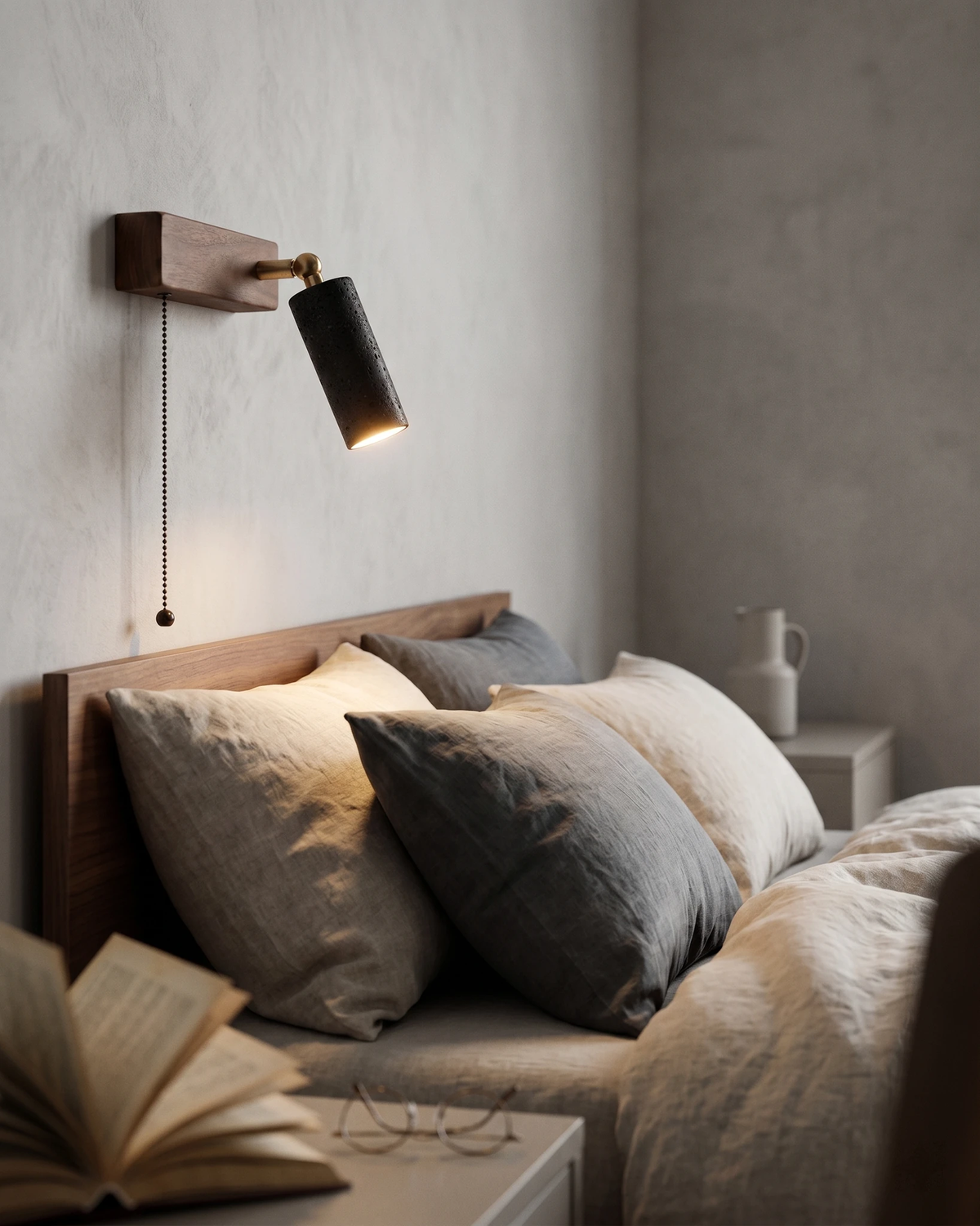 Travertine wall sconce VITRU No.01 in volcano black, lit on a plaster wall above a bed with gray pillows and a book