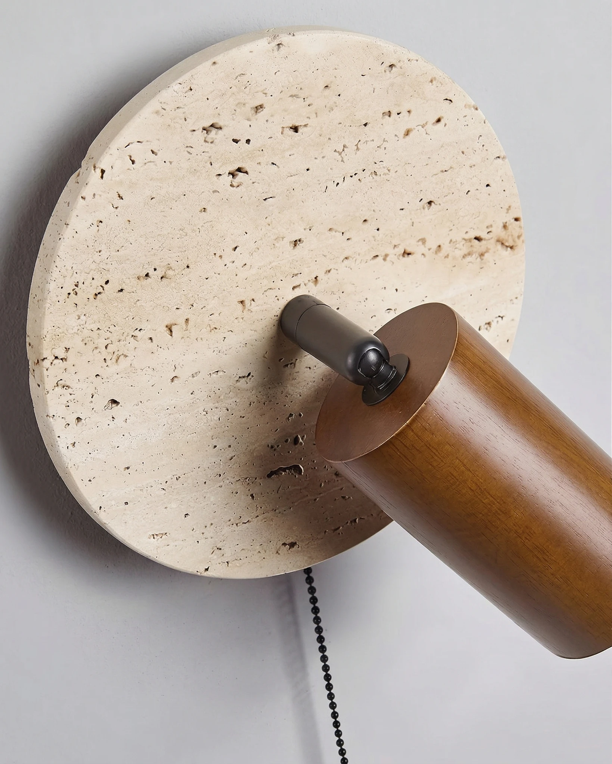Close-up of the travertine wall sconce VITRU No.03 showing the pitted stone disc, walnut cylinder and beaded pull chain