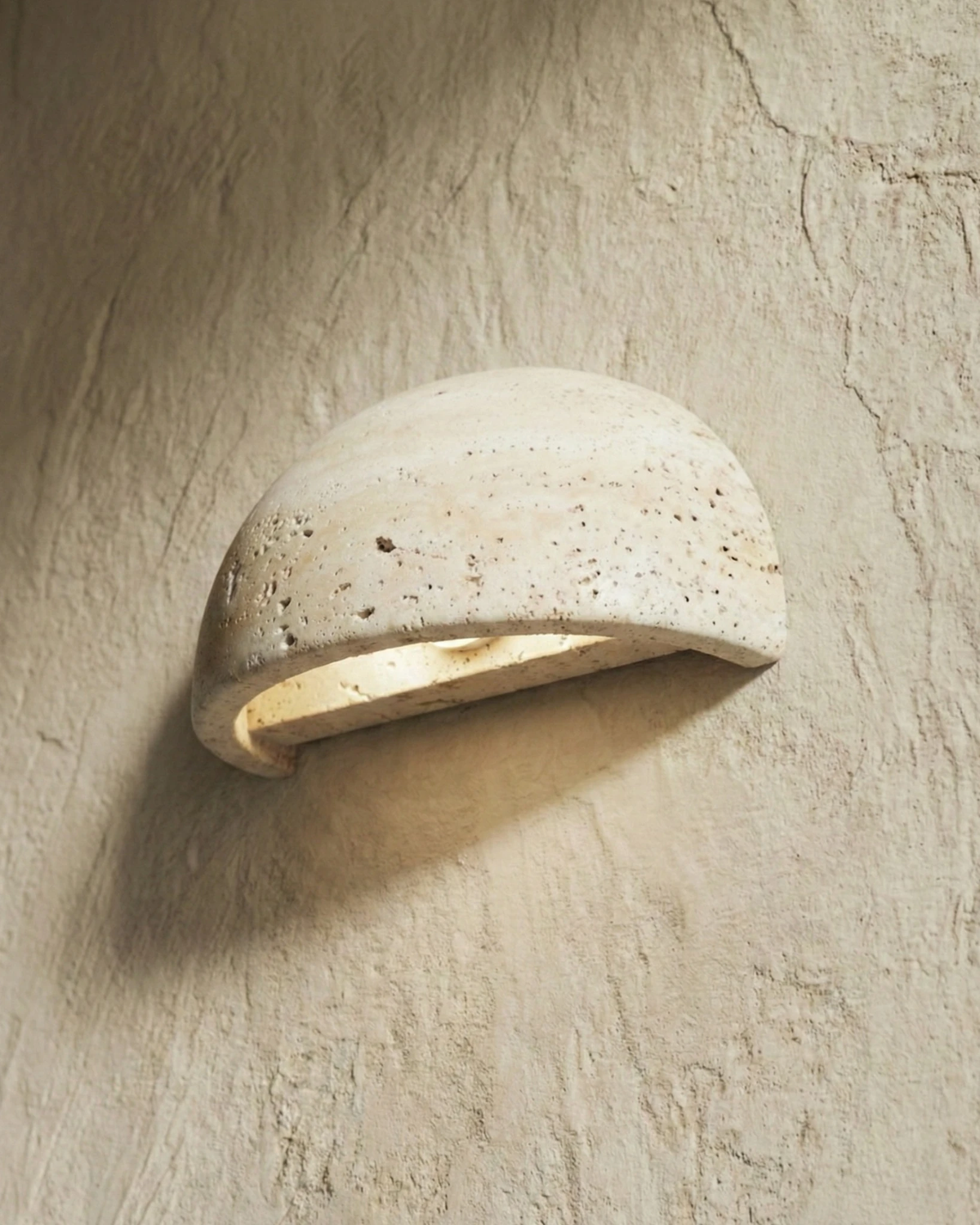 Close-up of the travertine wall sconce BERGAMO No.01, pitted yellow stone half-dome glowing against a plaster wall