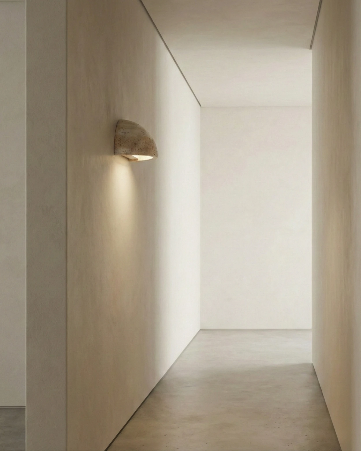 Travertine wall sconce BERGAMO No.01 switched on in a bare plaster hallway, casting a soft pool of light down the wall