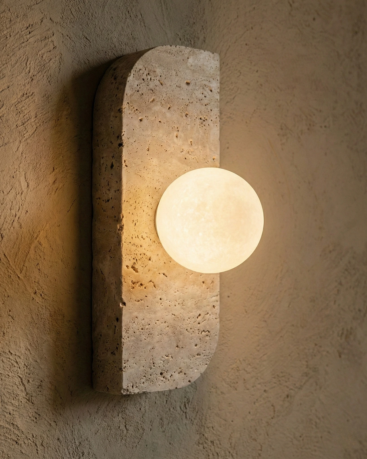 Travertine wall sconce GENOVA No.01 close view lit, pitted travertine plate and opal globe on a plaster wall