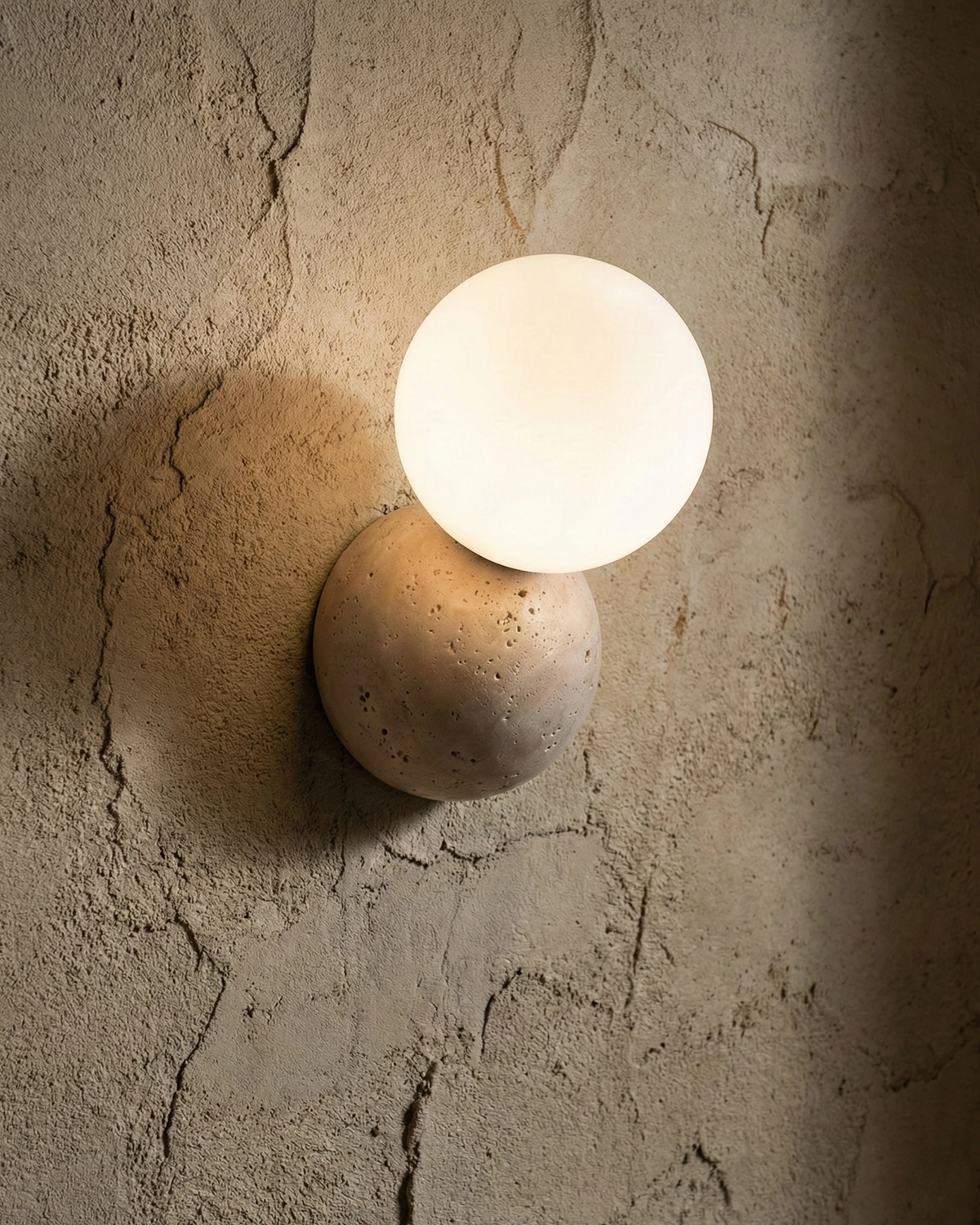 Travertine wall sconce MARE No.01 lit on a cracked plaster wall, glass globe glowing on a travertine disc