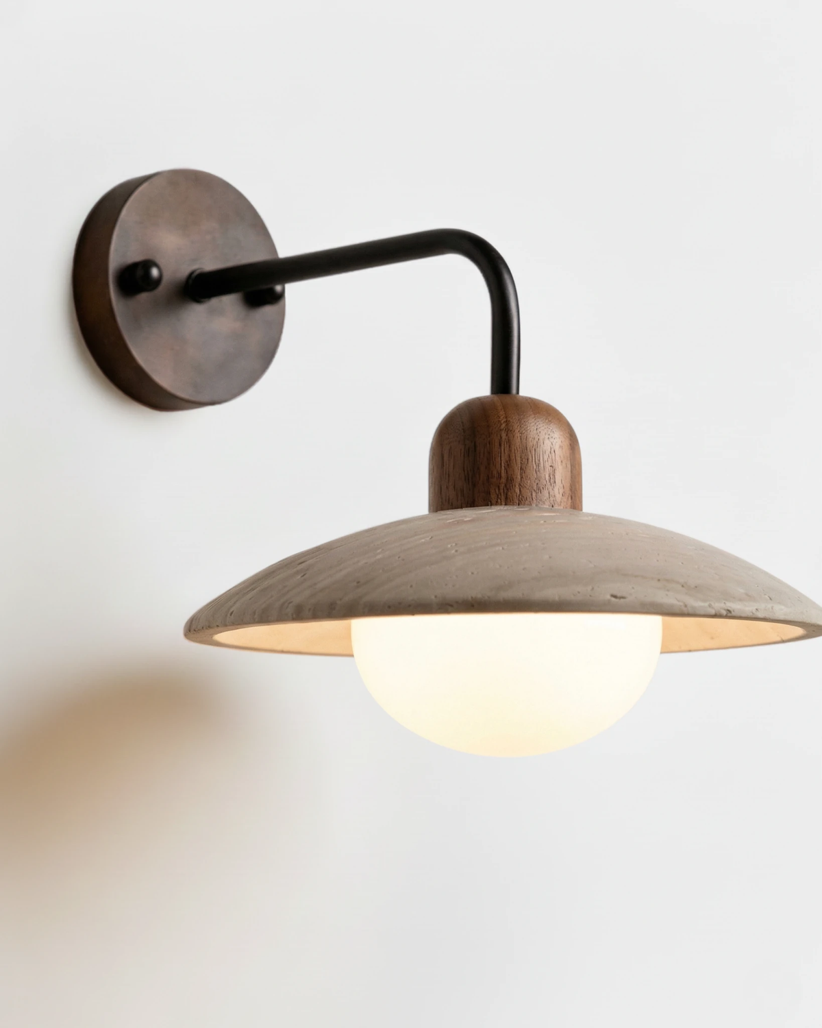 Travertine wall sconce IVERA No.01 with walnut cap and black arm, opal globe glowing beneath the stone disc