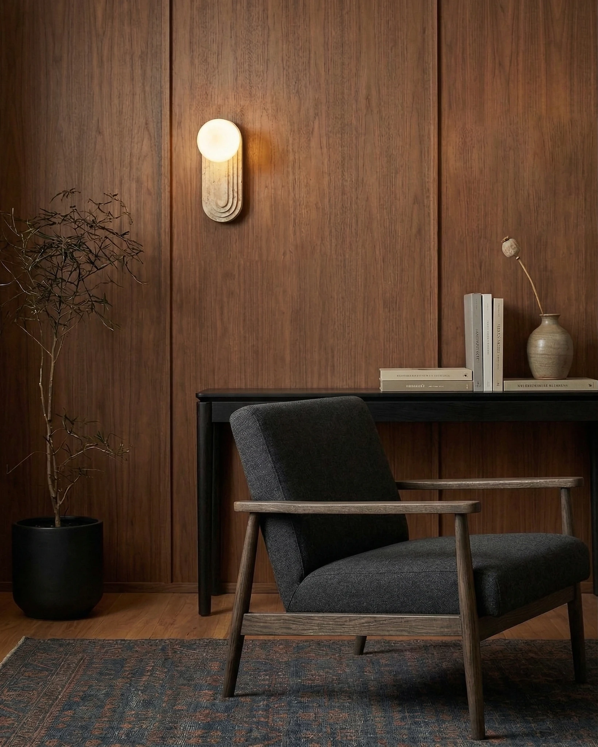 Travertine wall sconce ARCA No.01 lit on a walnut paneled wall above a black console and grey armchair