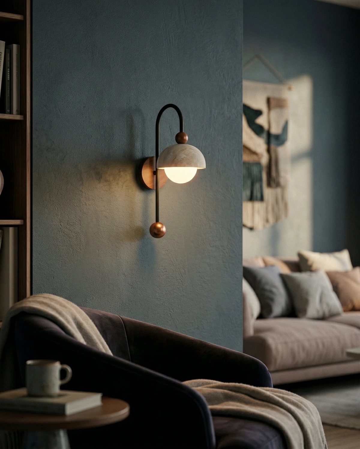 Travertine wall sconce IBLA No.01 lit on a dark blue wall above a navy armchair with a mug on a side table
