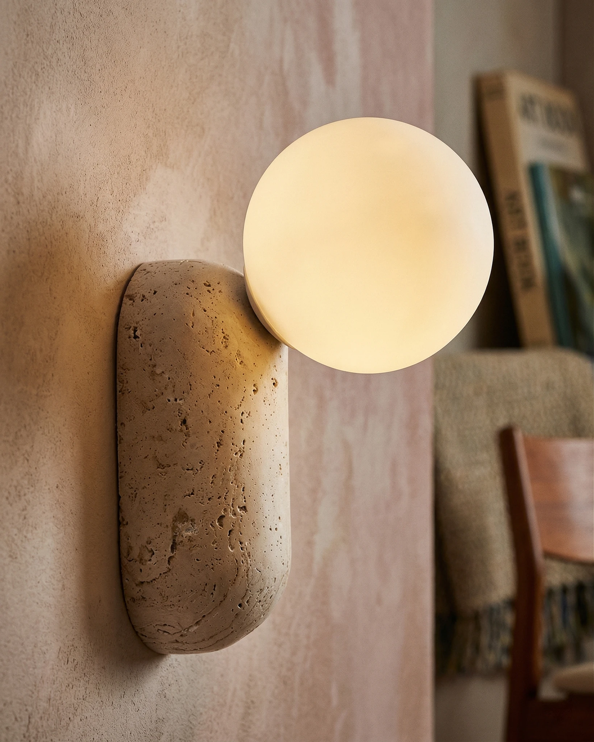 Travertine wall sconce PERLA No.01 lit on a pink plaster wall, opal globe on a raw travertine block, chair behind