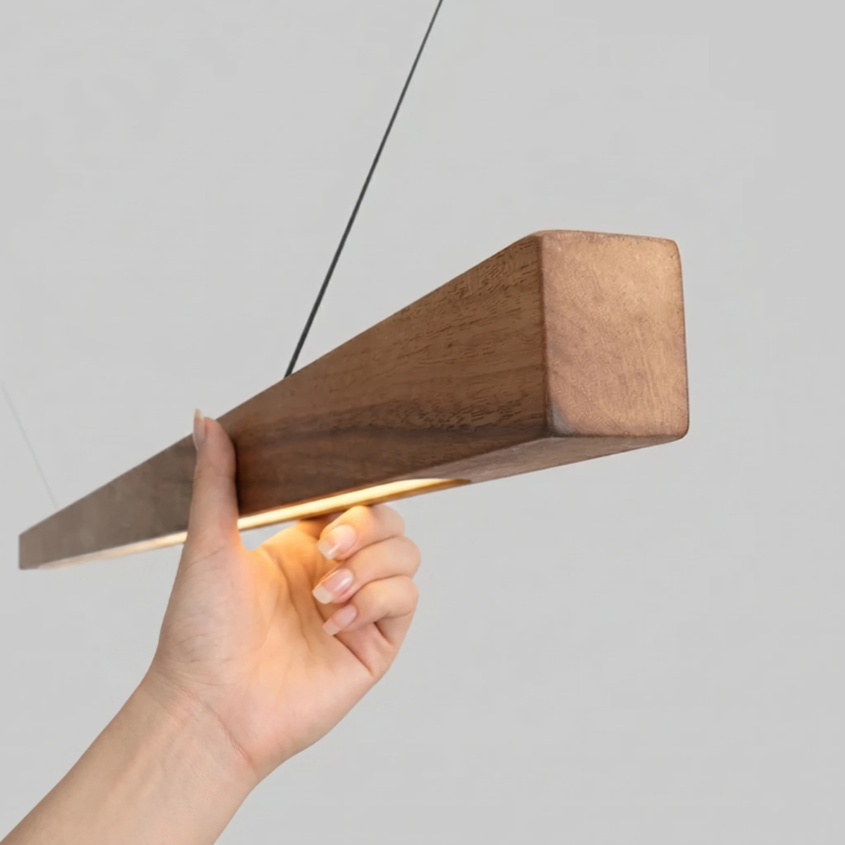 Close-up of the wood pendant light VIRGA No.01, a hand holding the walnut bar to show the lit LED strip on its underside