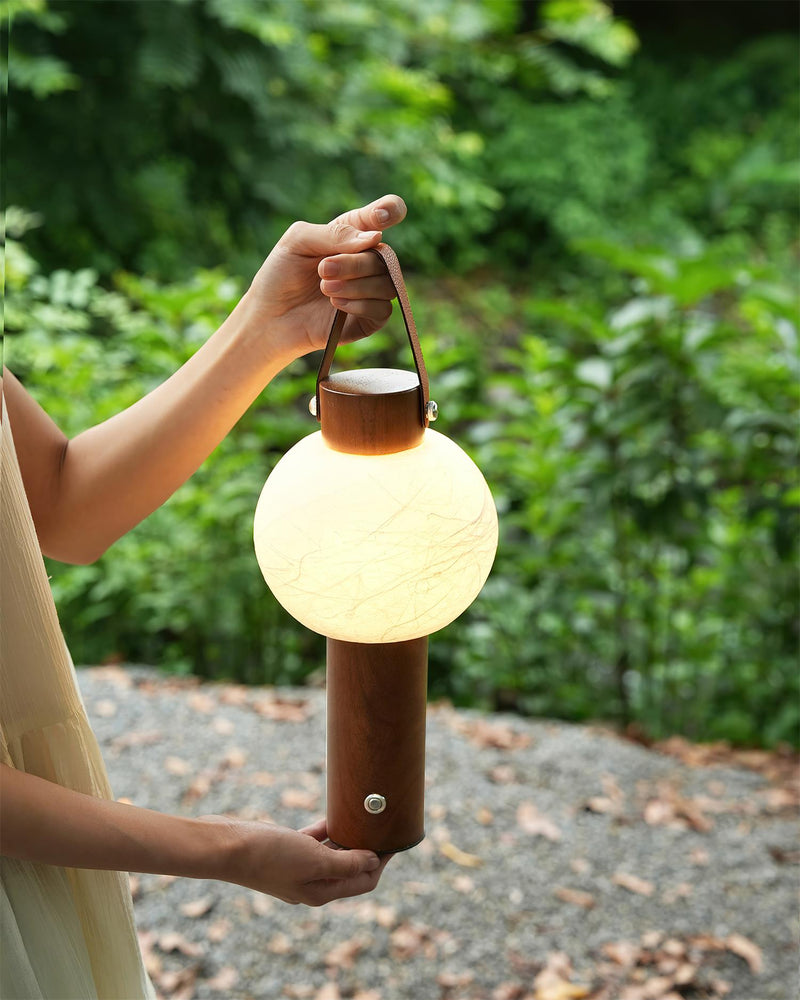 Wood table lamp NAPOLI No.01 carried outdoors by a woman in white, globe lit against dense greenery