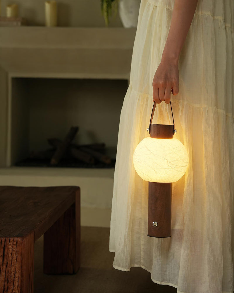 Wood table lamp NAPOLI No.01 lit and held by the strap handle beside a wooden bench in a plaster walled room