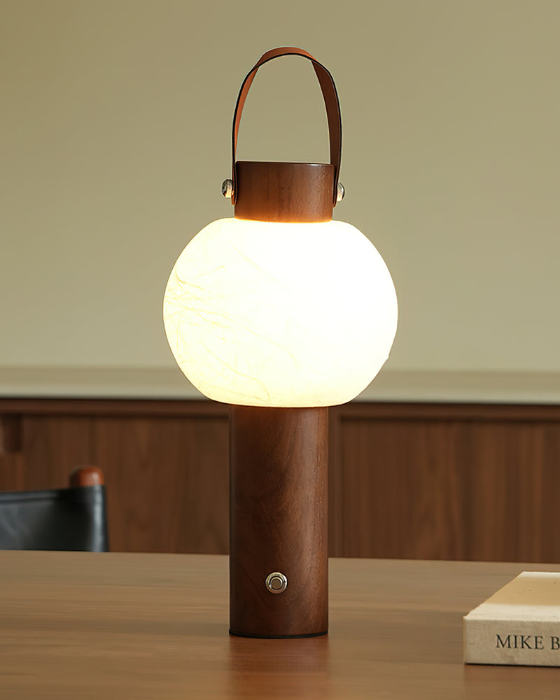 Wood table lamp NAPOLI No.01 in walnut lit on a wooden table beside a book, glowing globe with a leather handle