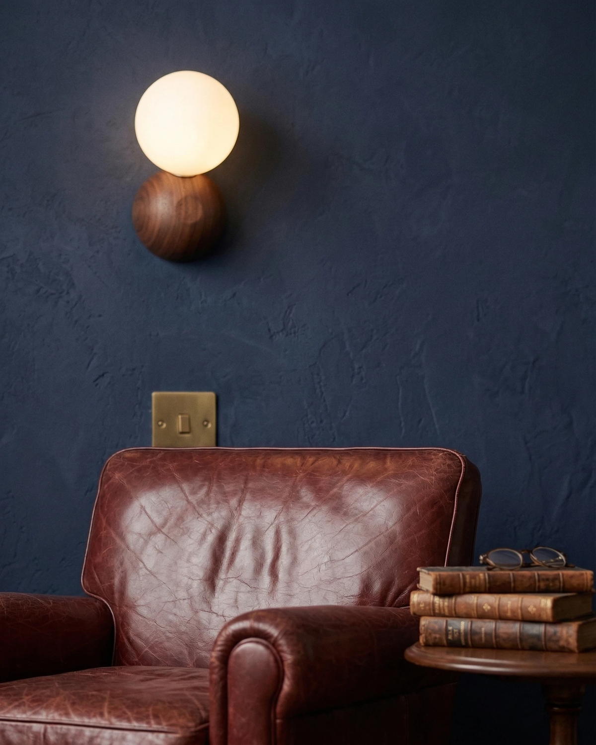 Wood wall sconce ALBE No.01, walnut pebble with a lit opal globe on a navy plaster wall above a leather armchair