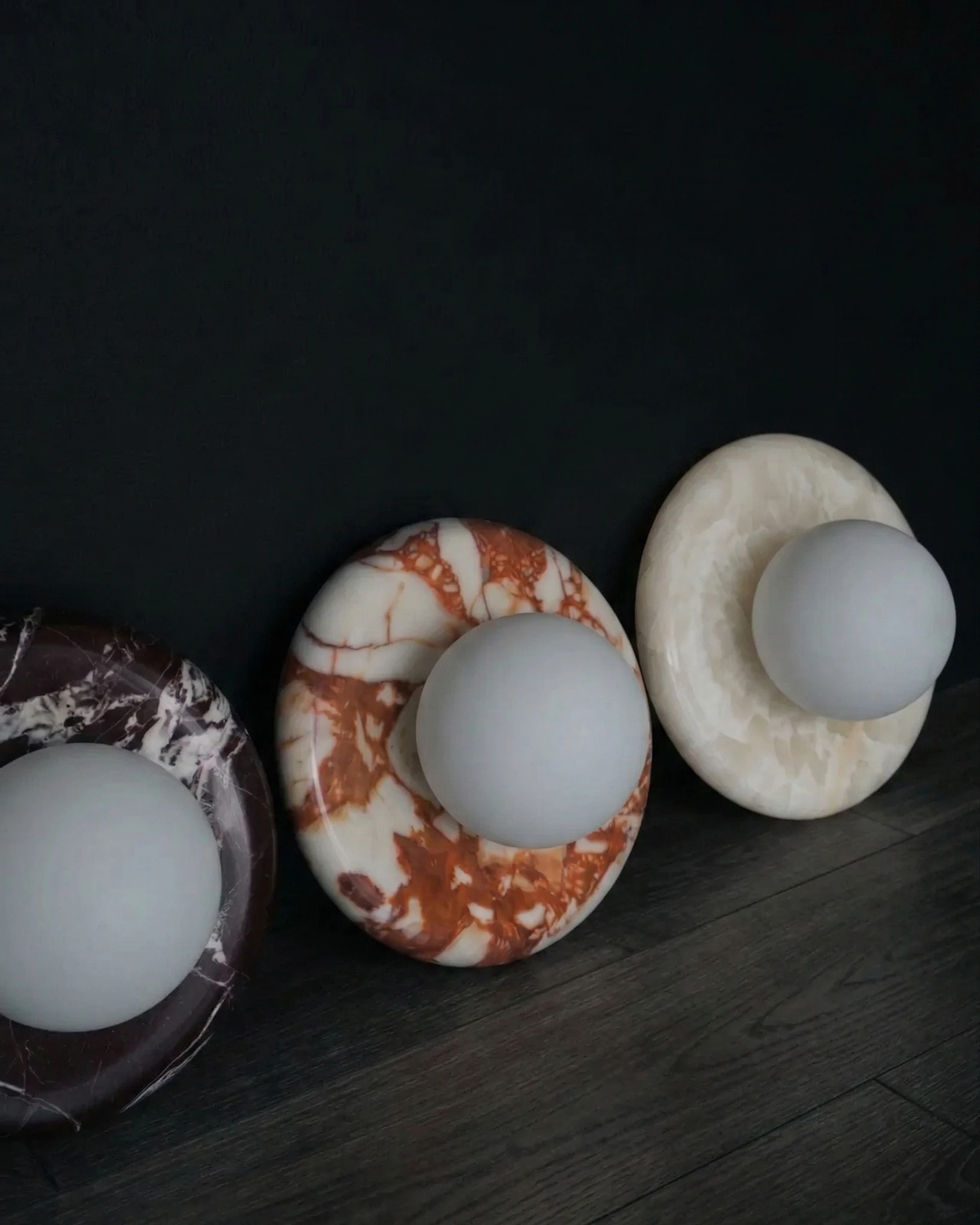 Three alabaster wall sconces LATTE No.02 in violet, red and white jade stone leaning against a dark wall, unlit