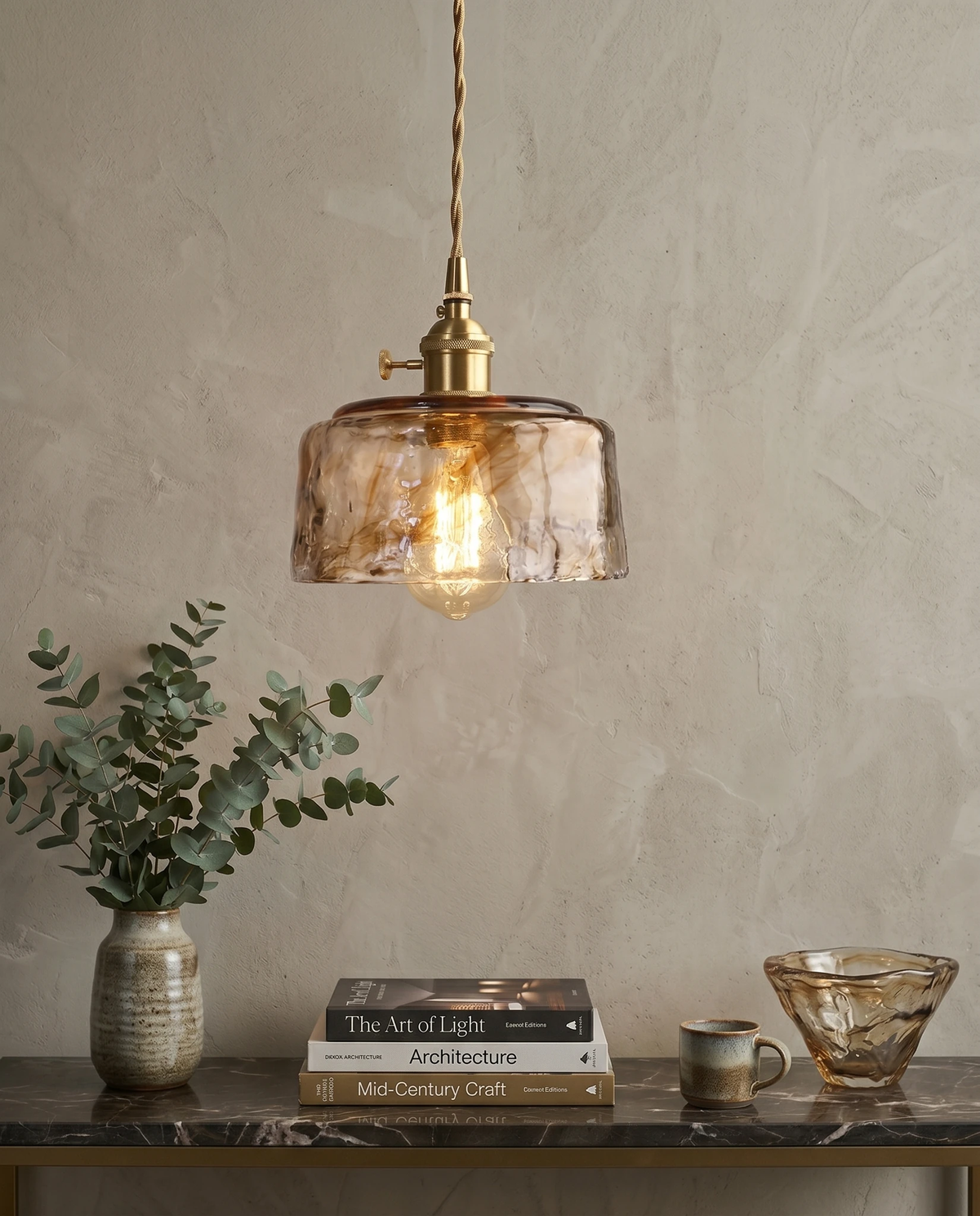 Amber glass pendant light OVIA No.05, Dome shade lit on a brass cord above a marble console with eucalyptus and books