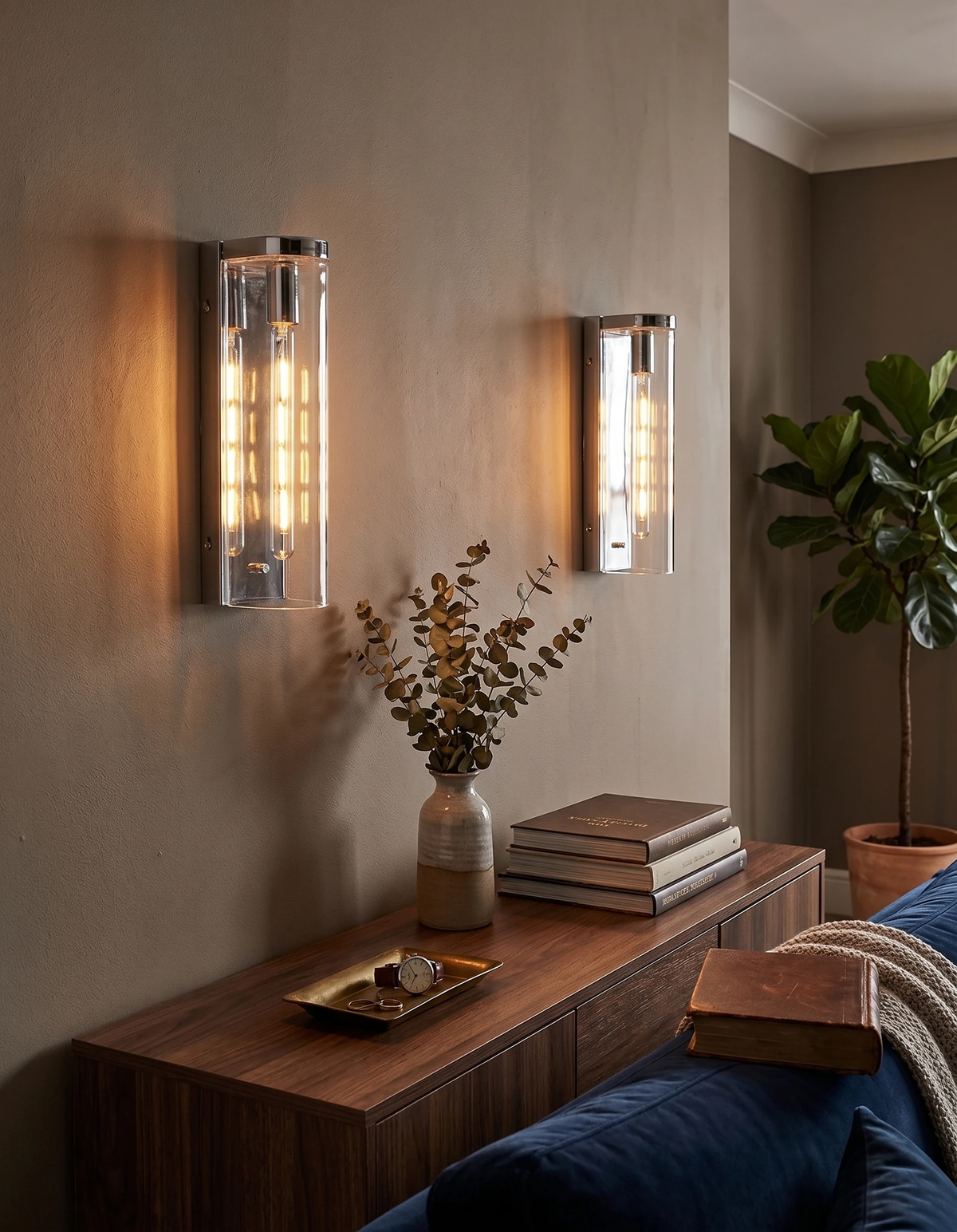 Brass wall sconce PILA No.01 in clear glass and polished chrome, a lit pair above a walnut console behind a navy sofa