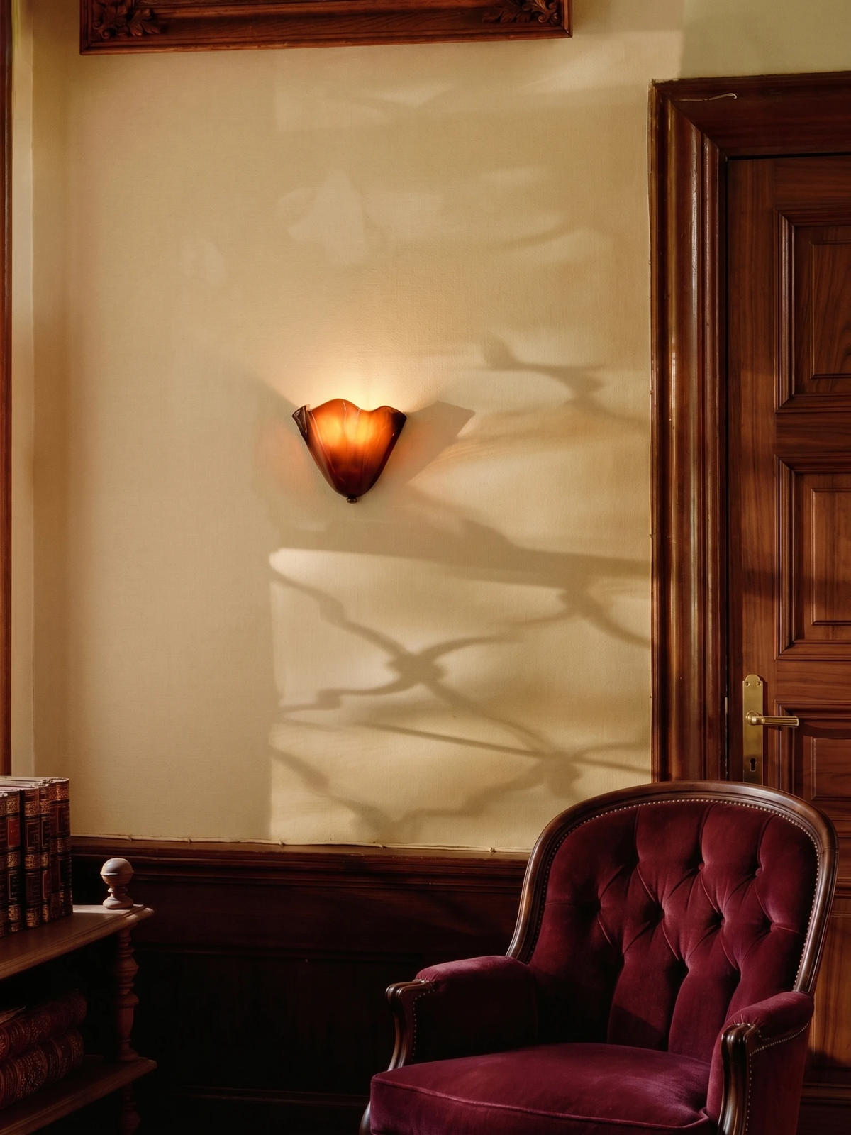 Brass wall sconce DALIA No.03 in amber glass lit on a cream wall beside a burgundy tufted armchair