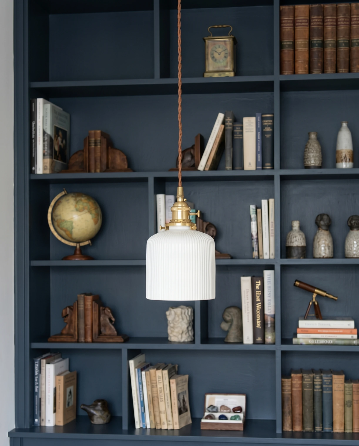 Ceramic pendant light SERA No.01 Fluted Cylinder lit in front of a navy blue bookcase filled with books and curios