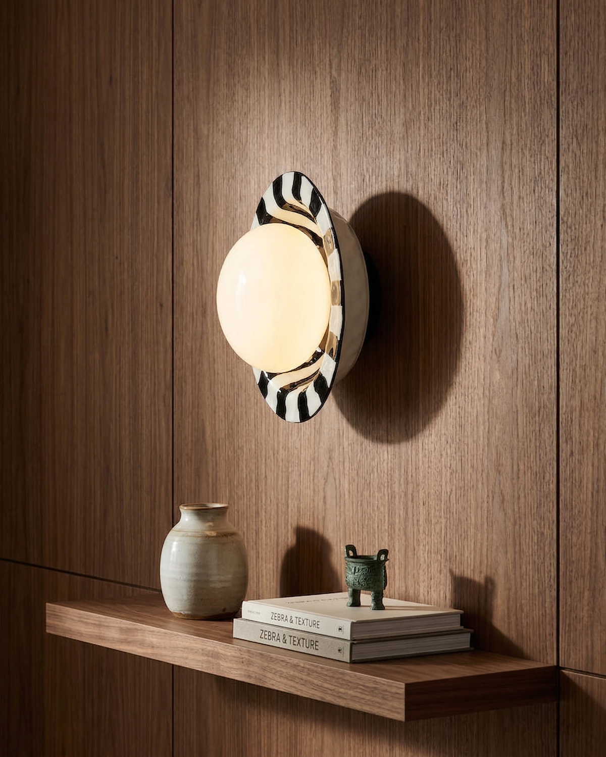 Ceramic wall sconce OLEA No.01 switched on against walnut paneling above a floating shelf with a vase and books