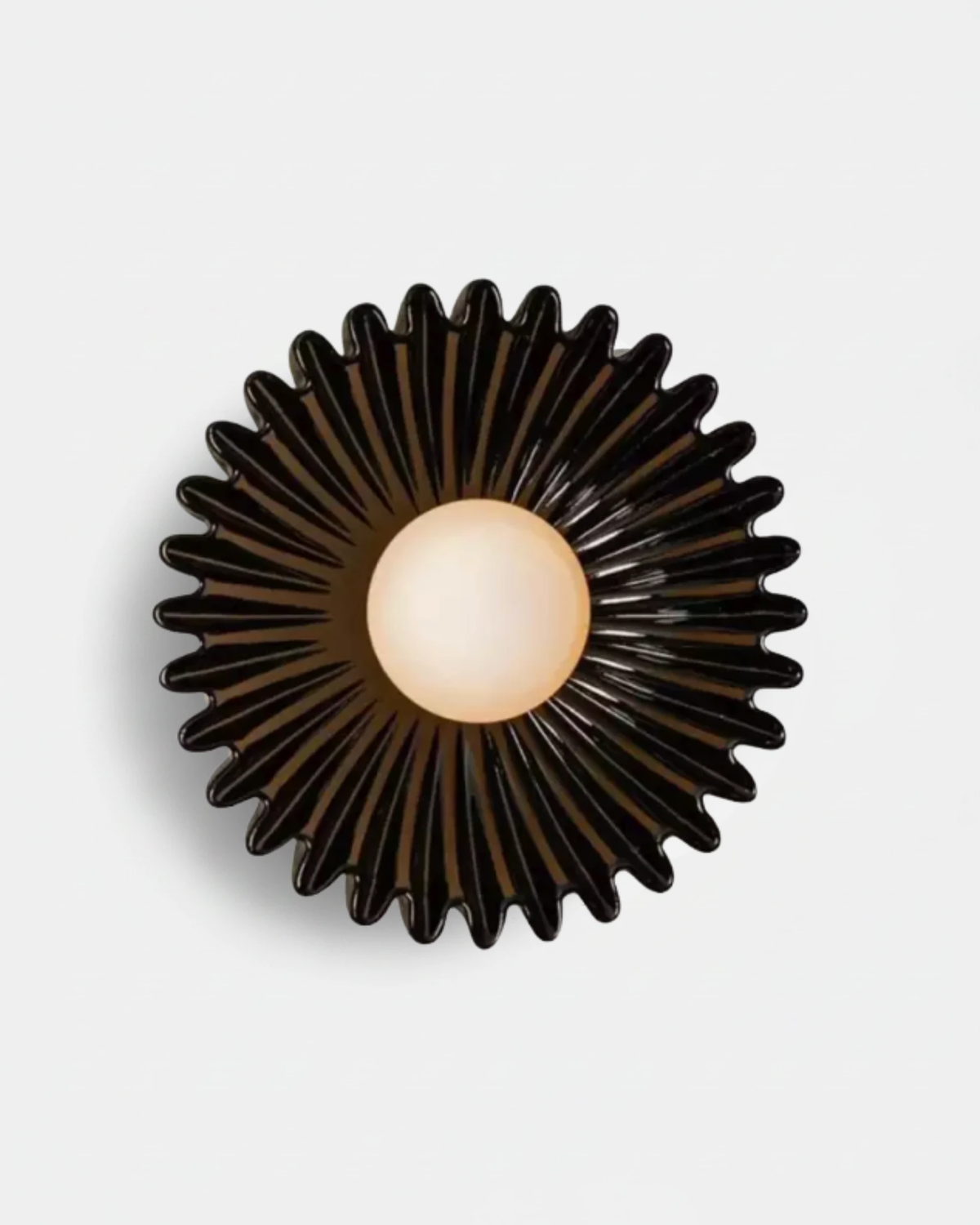 Ceramic wall sconce RAGGI No.01 in Volcano Black, glossy black ribbed sunburst with the opal globe glowing warm