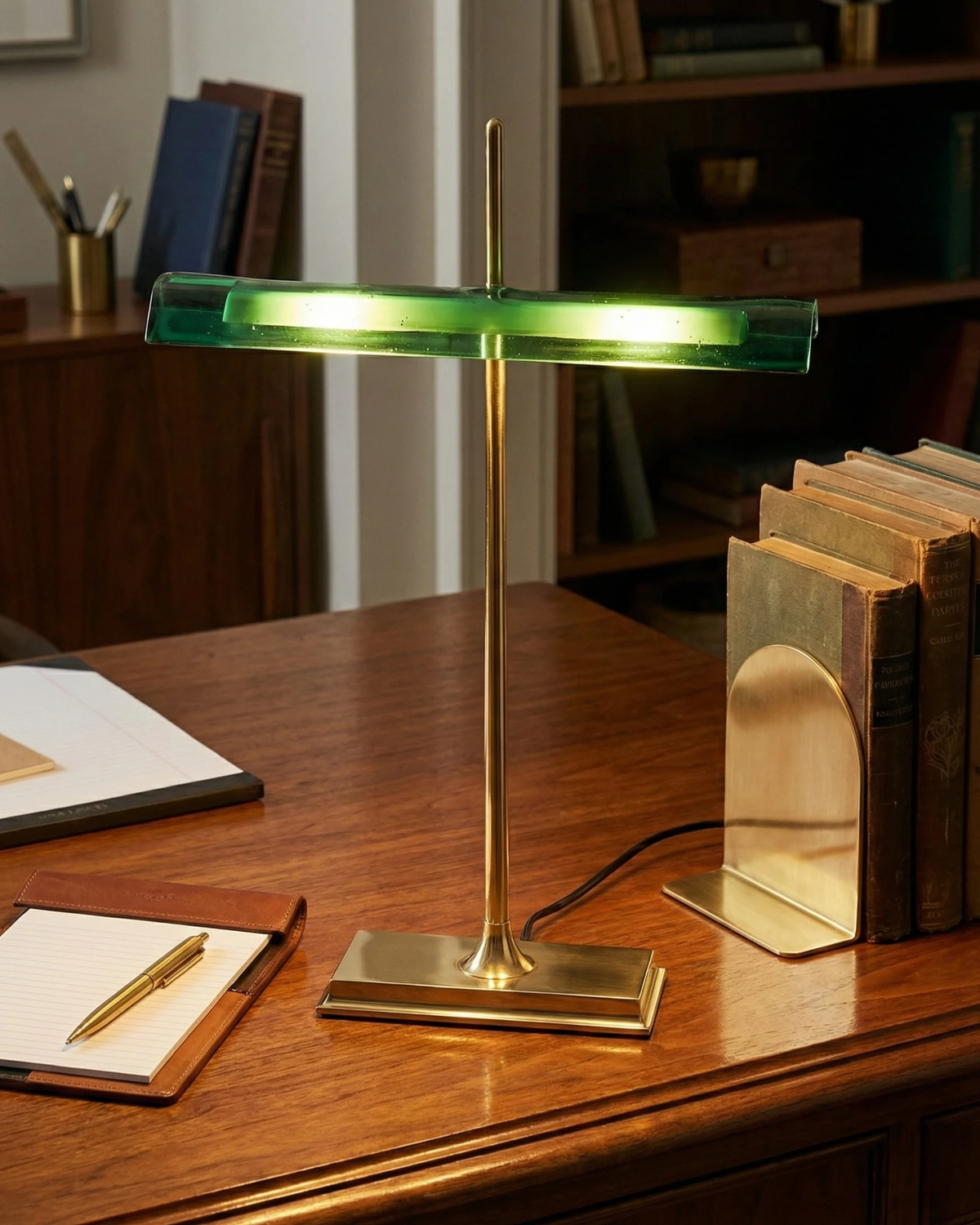 Cordless desk lamp BANCO No.02 in green lit on a mahogany desk with books, a notebook and a brass bookend