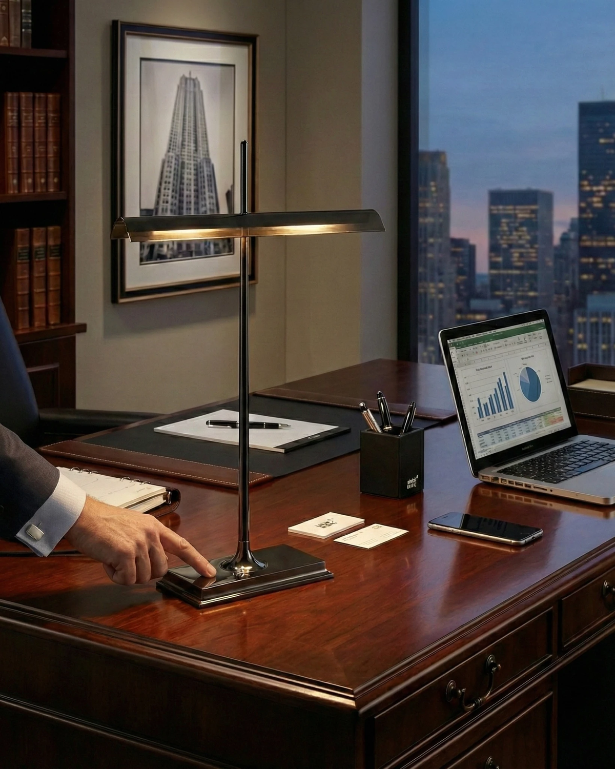 Cordless desk lamp BANCO No.02 in black lit on a wood desk with a laptop, a hand touching the base, city view behind