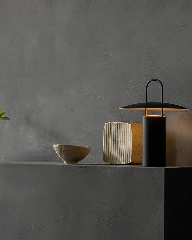 Cordless lantern lamp PORTO No.01 in black lit on a grey plaster ledge beside a bowl and a ribbed vase