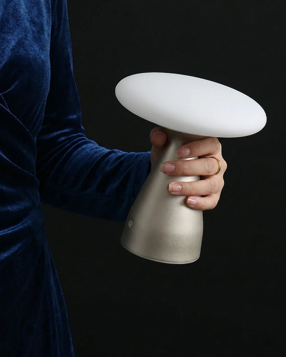 Cordless mushroom lamp DOMA No.03 held lit in one hand by a person in a blue velvet sleeve, dark background