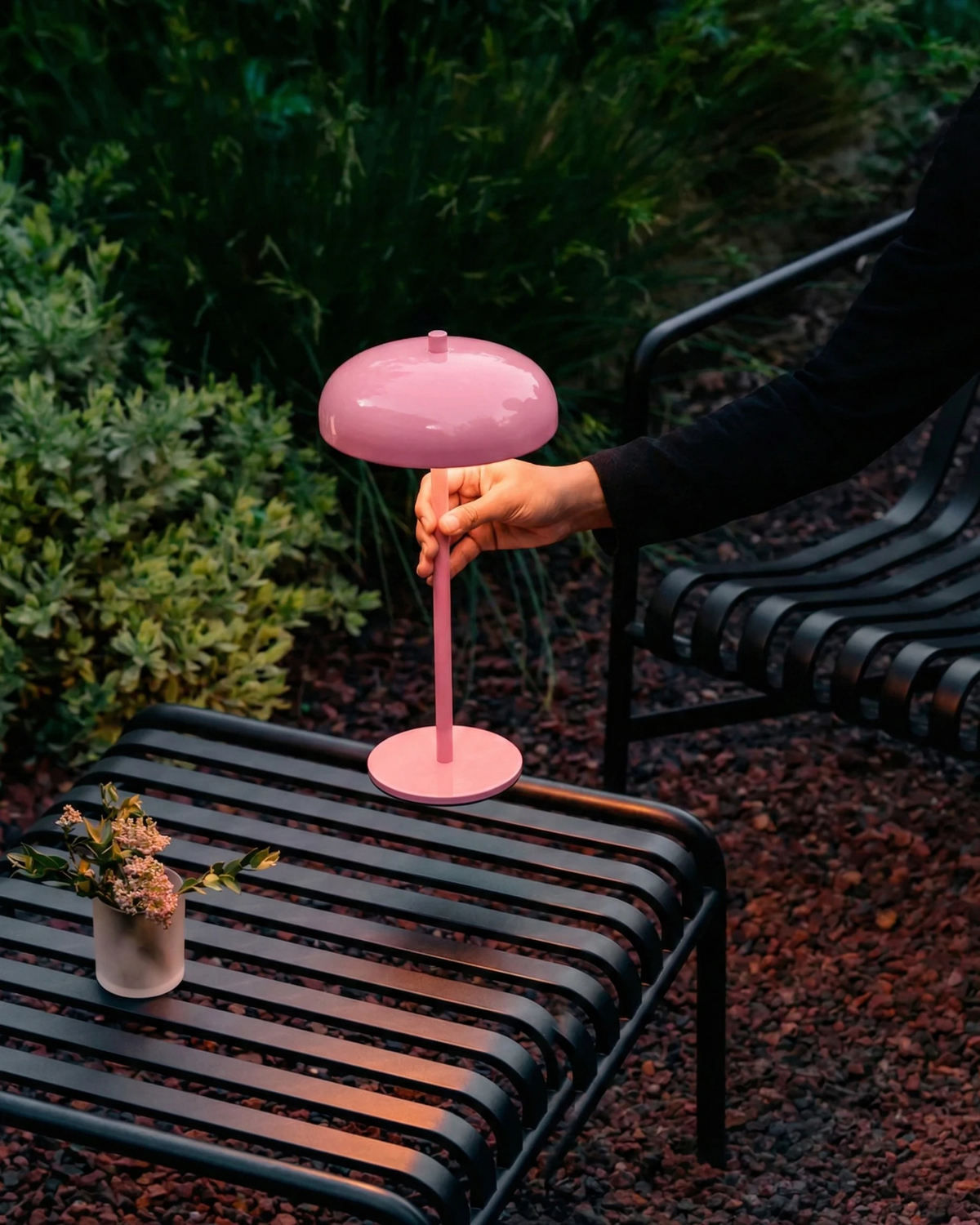 Cordless mushroom lamp DOMA No.01 in pink on a black slatted garden bench at dusk, a hand adjusting the shade