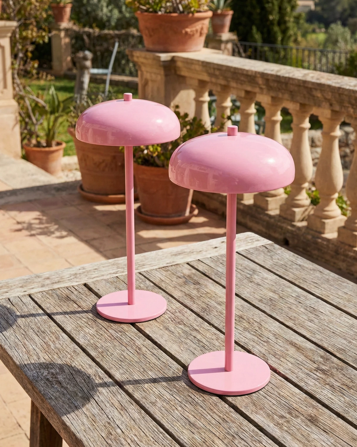 Two cordless mushroom lamps DOMA No.01 in pink on a teak outdoor table on a stone terrace with potted plants