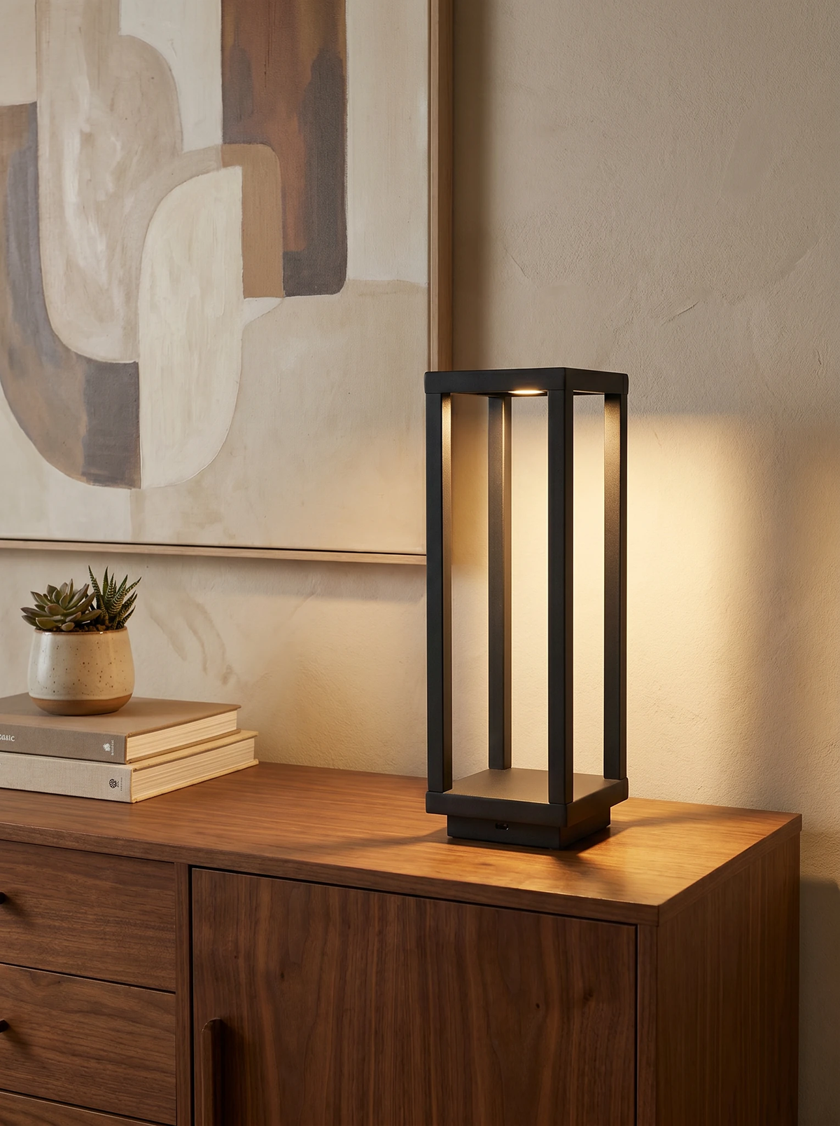 Cordless outdoor table lamp FARO No.07, black open-frame lantern lit on a walnut sideboard below abstract art