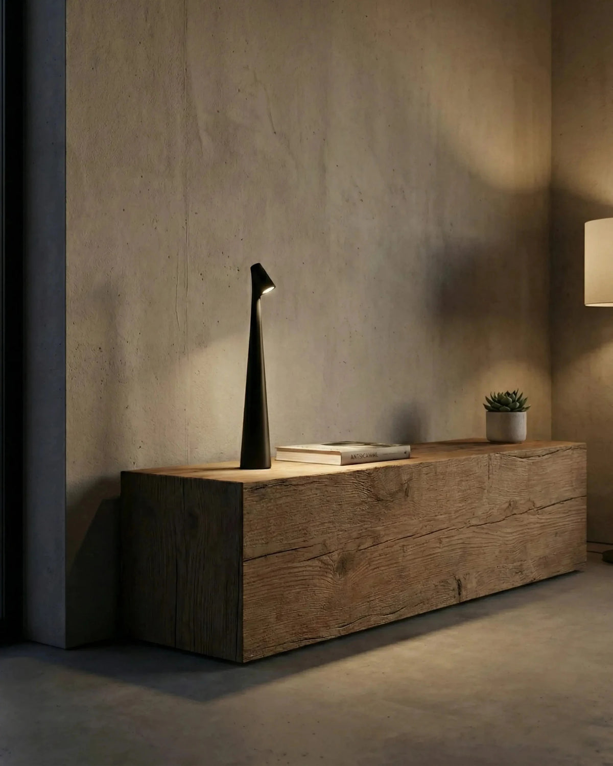 Cordless reading lamp TRIESTE No.01 in black switched on atop a rustic wood block against a concrete wall