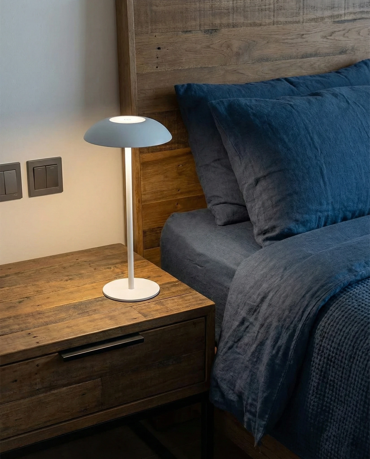 Cordless table lamp PESCARA No.01 in white lit on a rustic wood nightstand next to a bed with blue linen