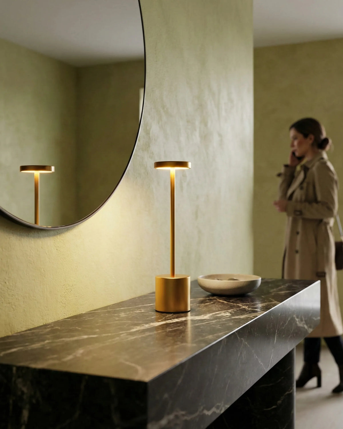 Cordless table lamp RIETI No.01 in brushed gold, lit on a black marble console below a round mirror as a woman walks past