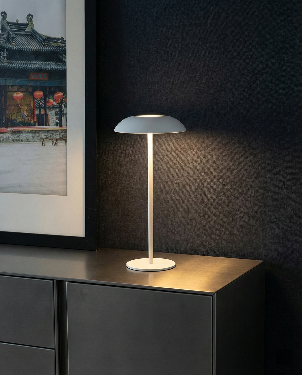 Cordless table lamp PESCARA No.01 in white lit on a dark metal sideboard beside a framed print