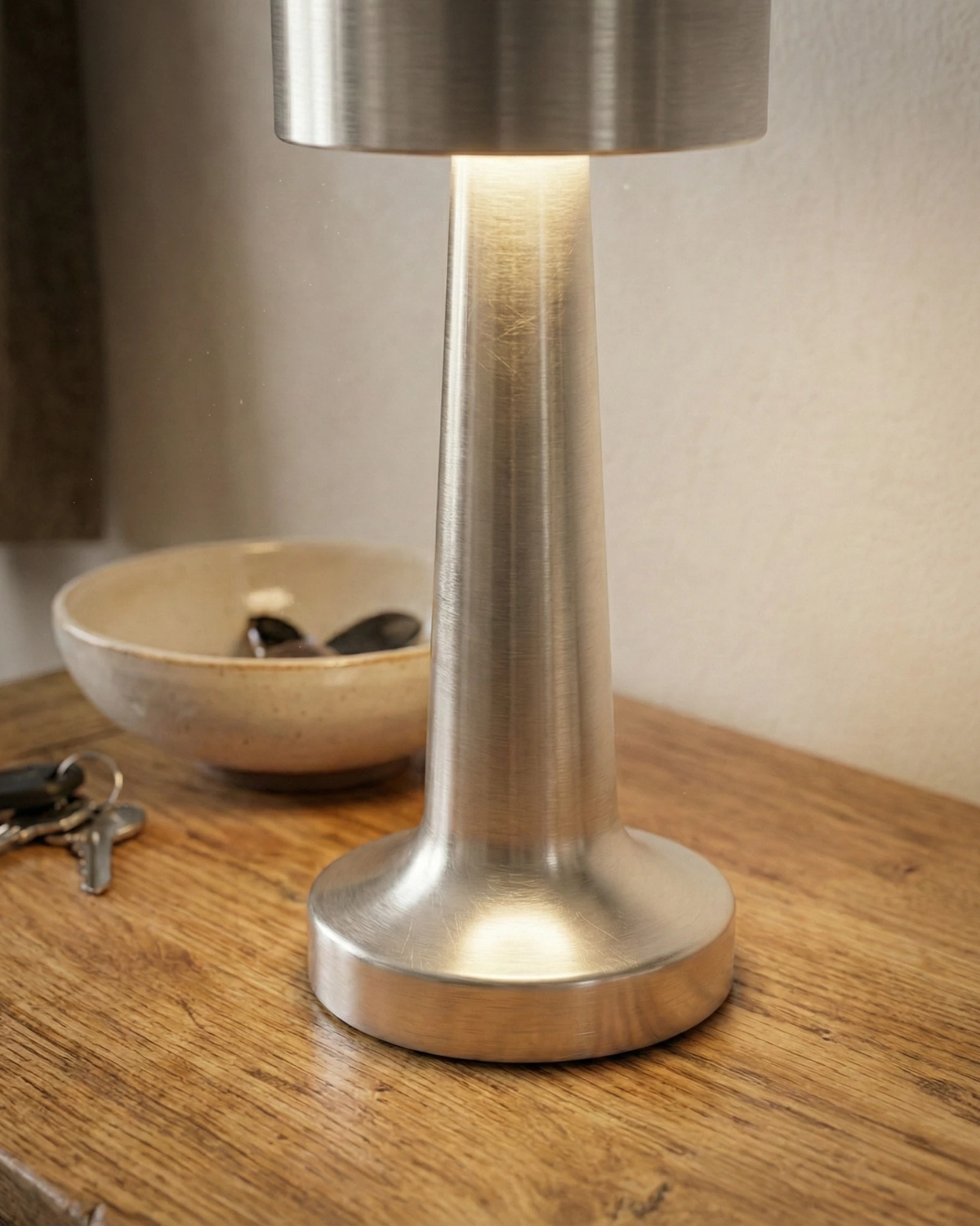 Close-up of the ISOLA No.01 cordless table lamp in silver lit on a wooden table beside a bowl